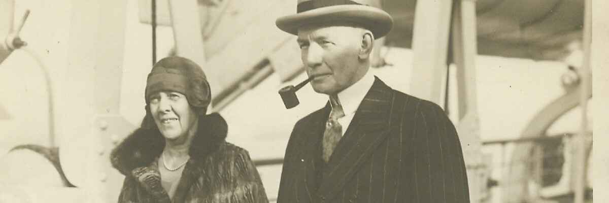 A horizontal, sepia-toned vintage photo of a man and a woman in 1920s-era attire. The man, on the right, wears a fedora, a dark suit, and a tie while smoking a pipe. The woman, on the left, wears a dark cloche hat and a patterned coat with a thick fur collar.