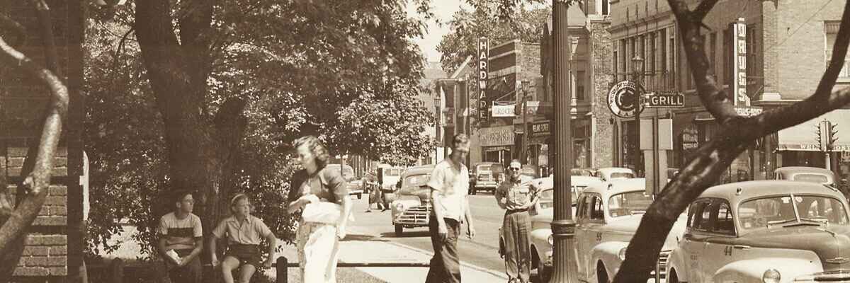 epia-toned historical photograph of a busy Evanston street scene with pedestrians walking along the sidewalk and vintage cars parked along the curb. Large leafy trees line the street, partially shading the storefronts and theater marquee in the background.