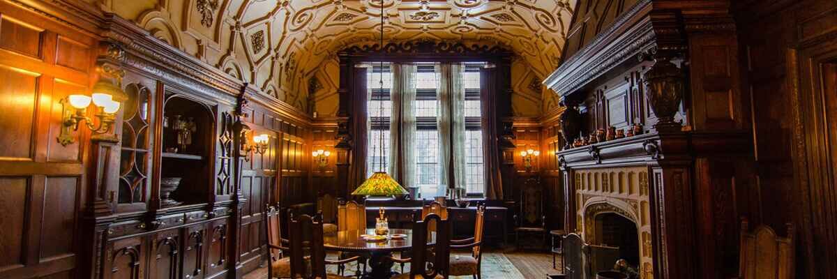 Interior view of a grand, wood-paneled dining room at the Dawes House featuring an ornate vaulted ceiling, a large stone fireplace to the right, and a central table with a green-shaded lamp positioned before a tall triptych window.